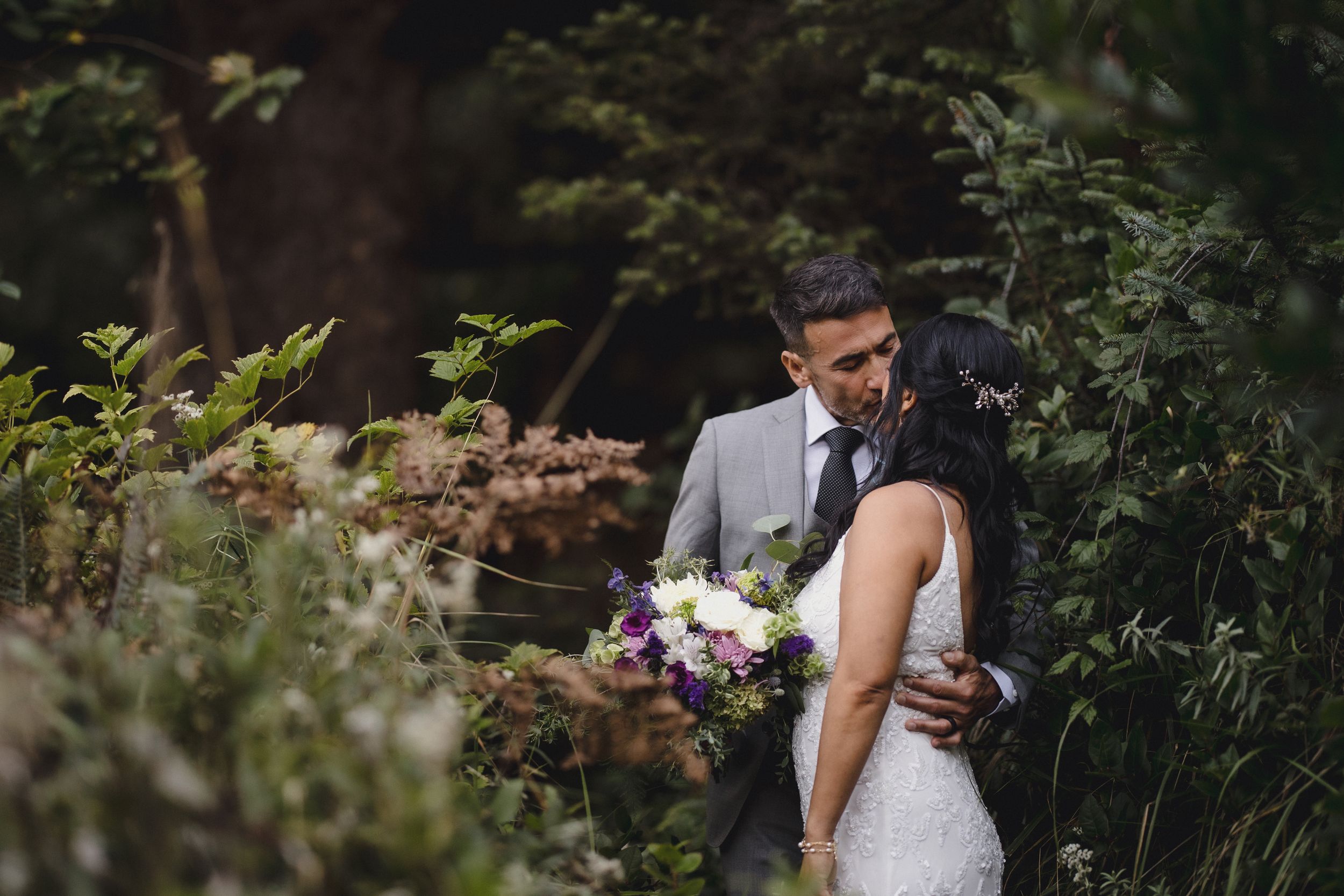 Bride and groom embrace in a lush garden, bride holding a purple and white bouquet in a romantic wedding portrait.