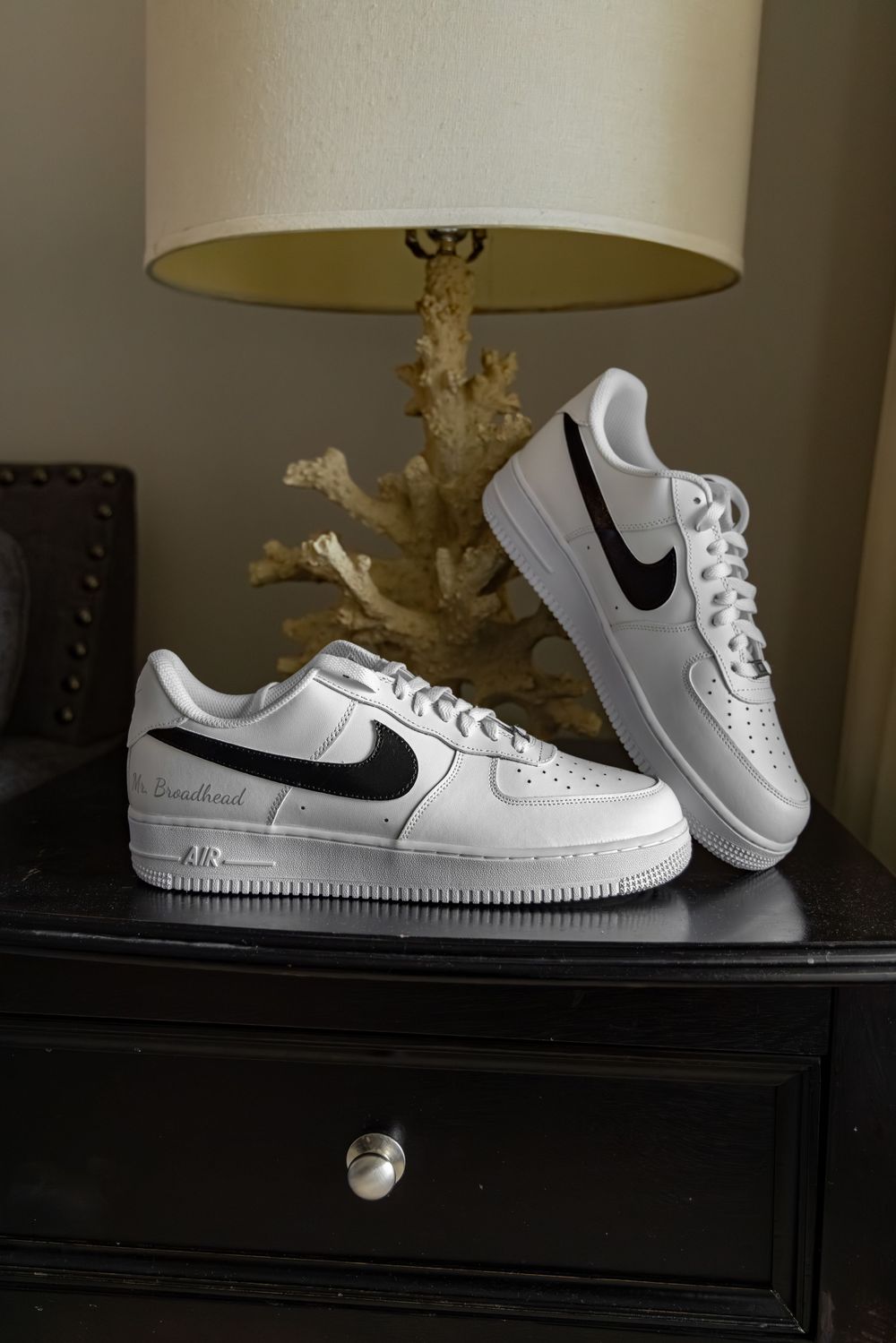White Nike Air Force 1 sneakers with black swoosh displayed on dark nightstand beside lamp.