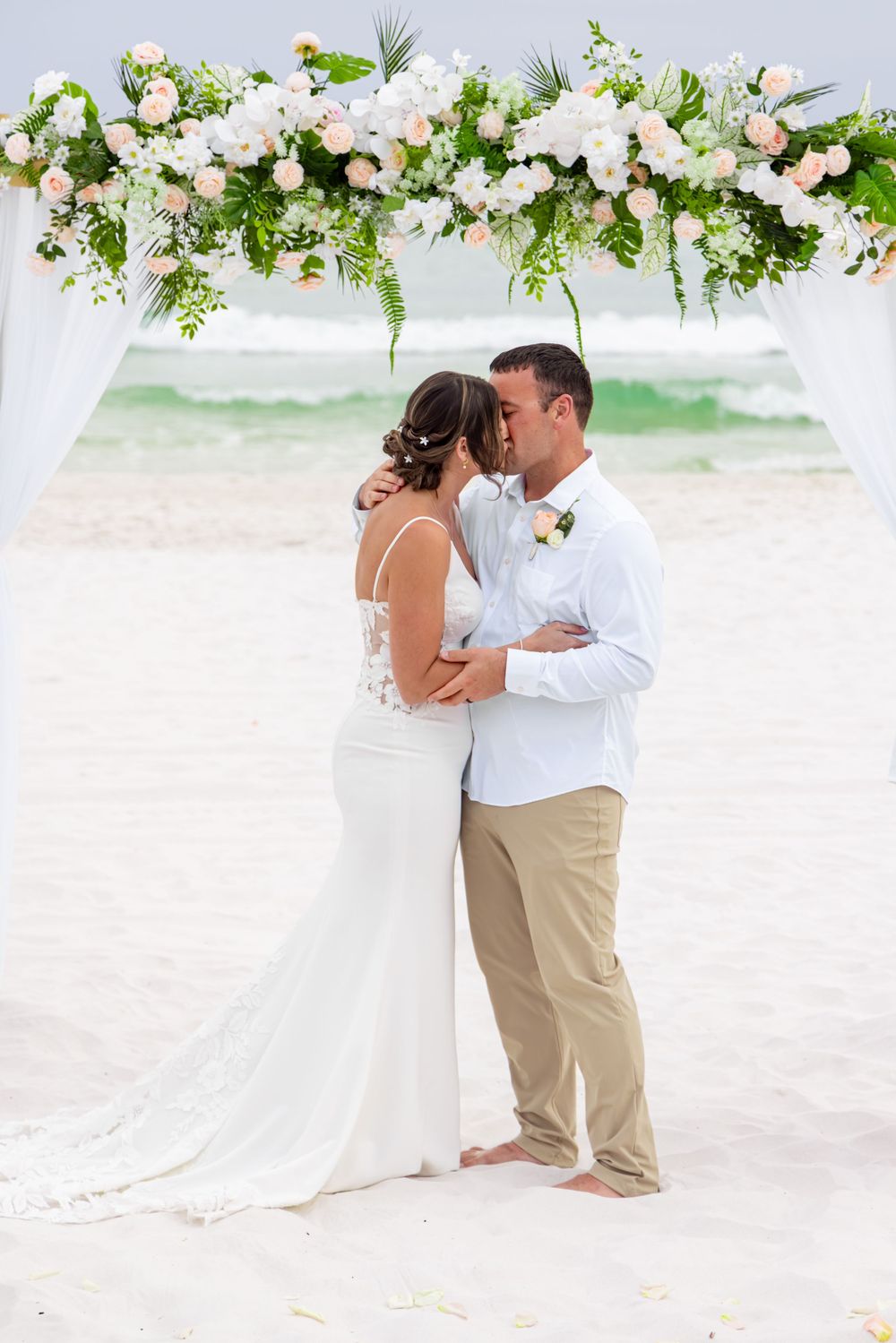 Bride and groom share first kiss at romantic beach wedding ceremony under floral arch with white and pink roses.
