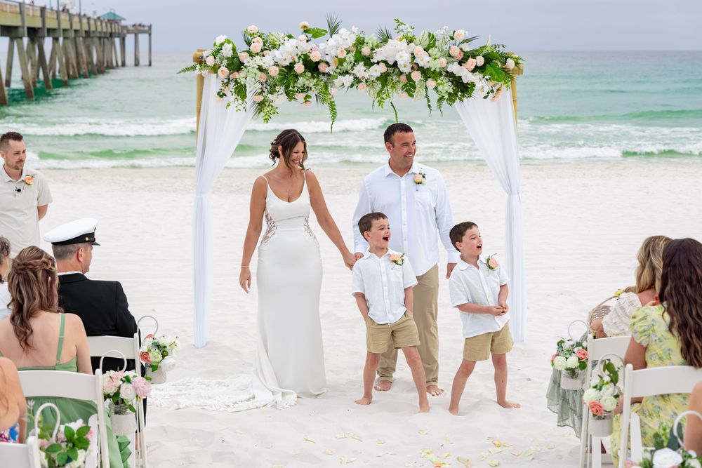 Bride and groom with two young boys at a beach wedding ceremony under a floral arch with white chairs and ocean backdrop.