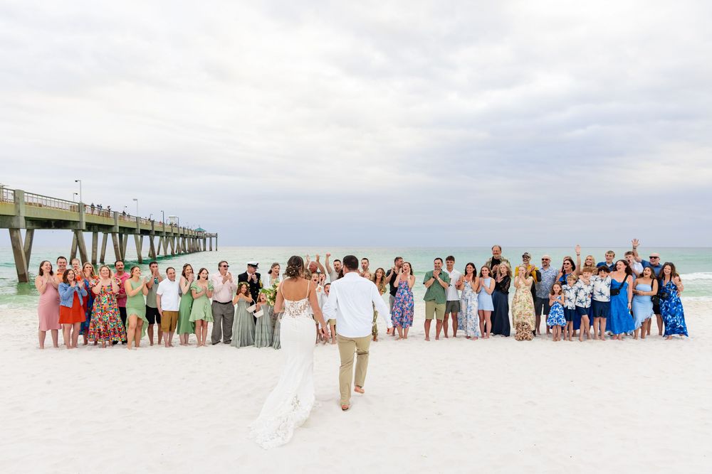 Bride and groom face wedding guests gathered on a white sand beach near a pier for a seaside ceremony.