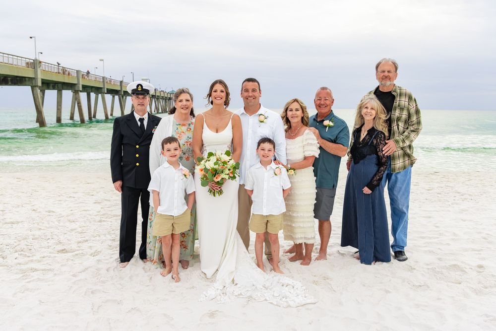 Beach wedding party poses on white sand near pier, bride in white gown with groom and family members.