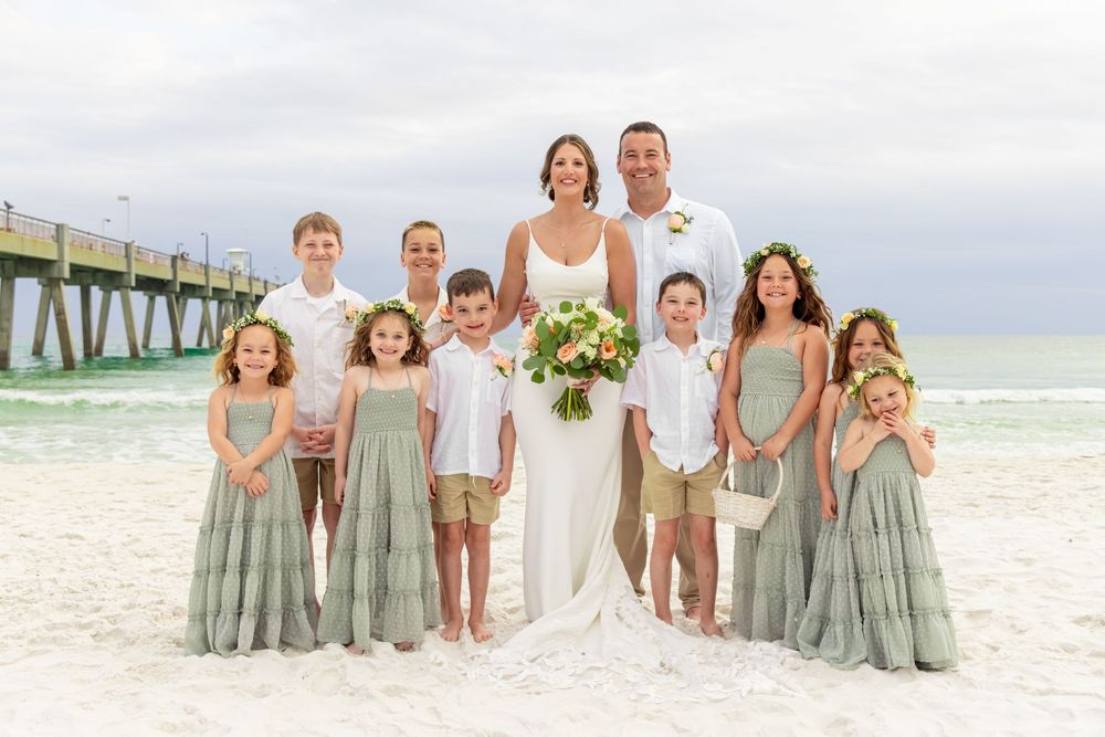 Beach wedding couple with children in sage green and white attire posing on sandy shore near pier.
