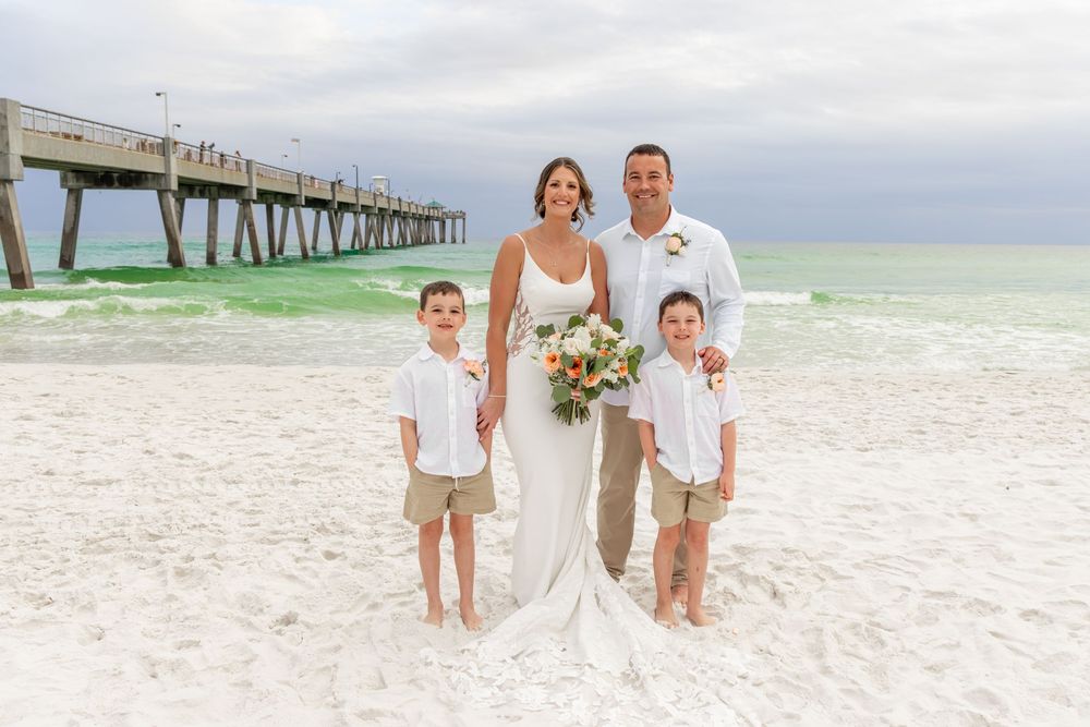 Beach wedding couple with two boys in white shirts posing on sandy shore near a pier.
