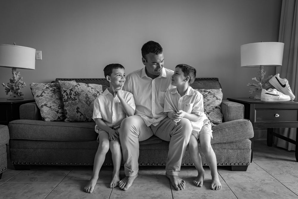 Black and white photo of a father sitting on a couch with his two young sons, sharing a warm family moment.