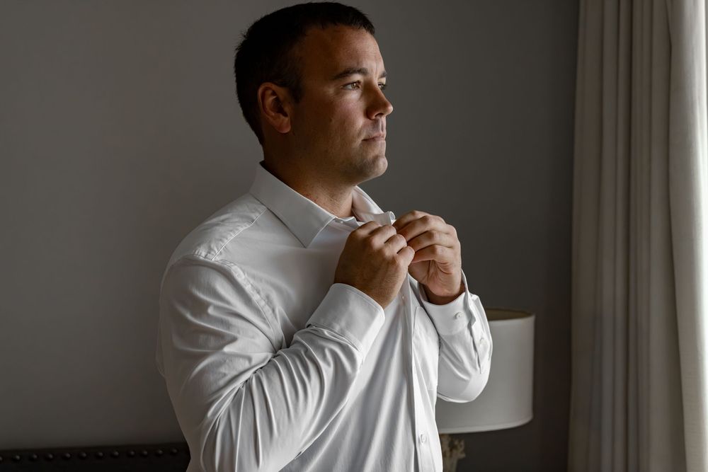 Man in white dress shirt buttoning his collar near a window, preparing for a formal event or wedding.