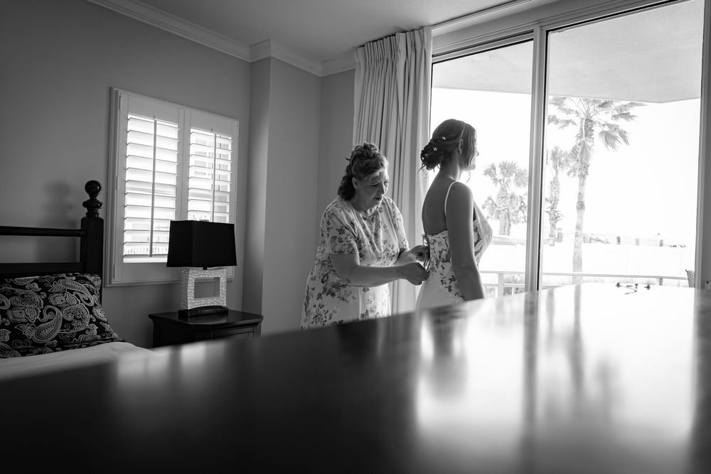 Black and white photo of two women in elegant dresses sharing a moment by a sunlit window on a wedding day.