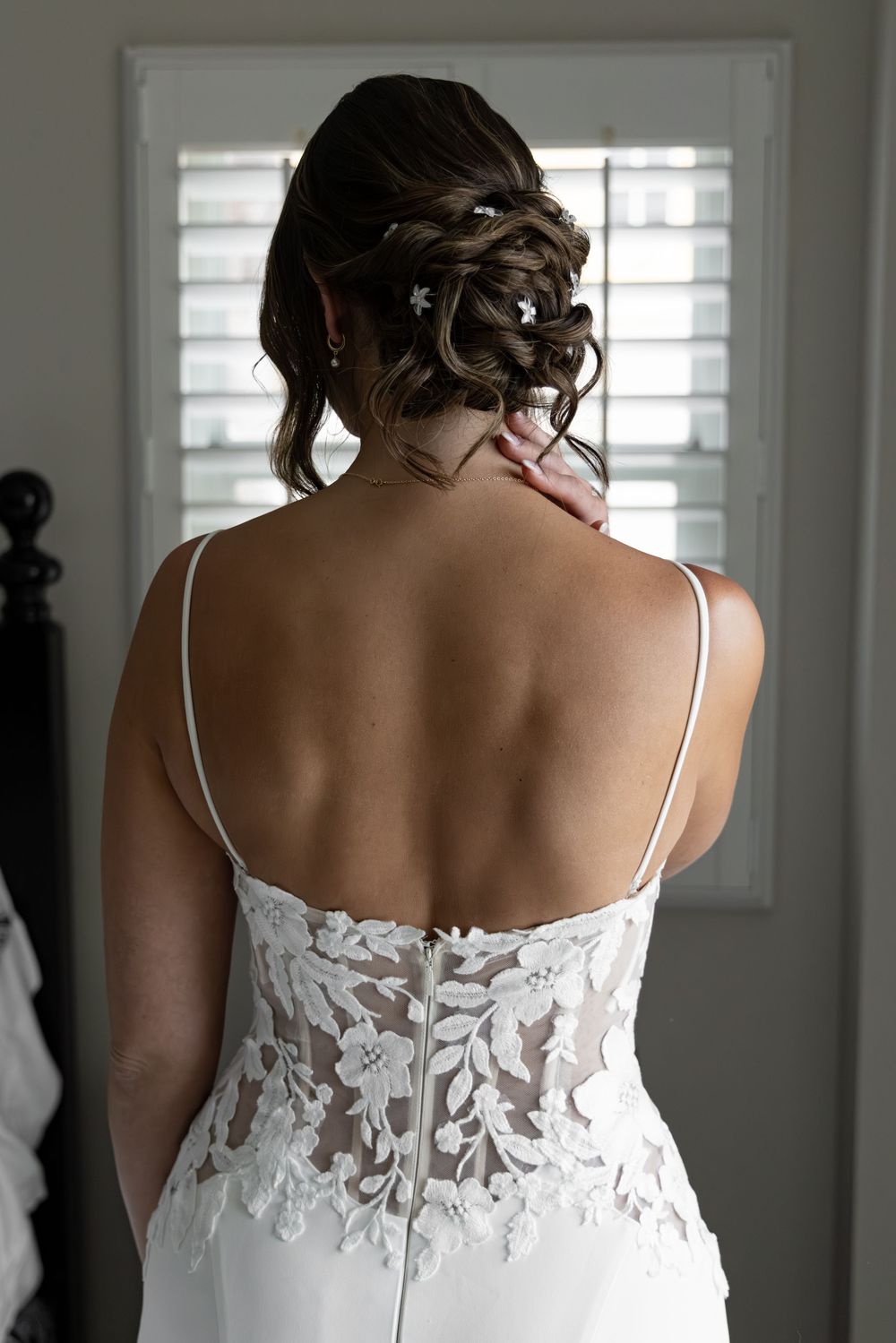 Bride in white lace-back wedding gown with spaghetti straps, hair in elegant updo with floral pins, viewed from behind.