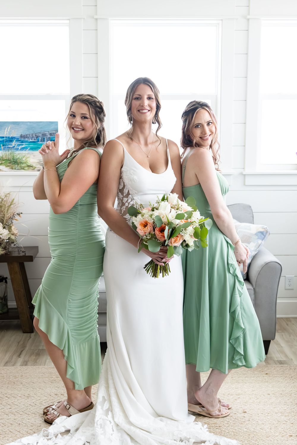 Bride in white gown holds bouquet flanked by two bridesmaids in sage green dresses posing indoors near bright windows.