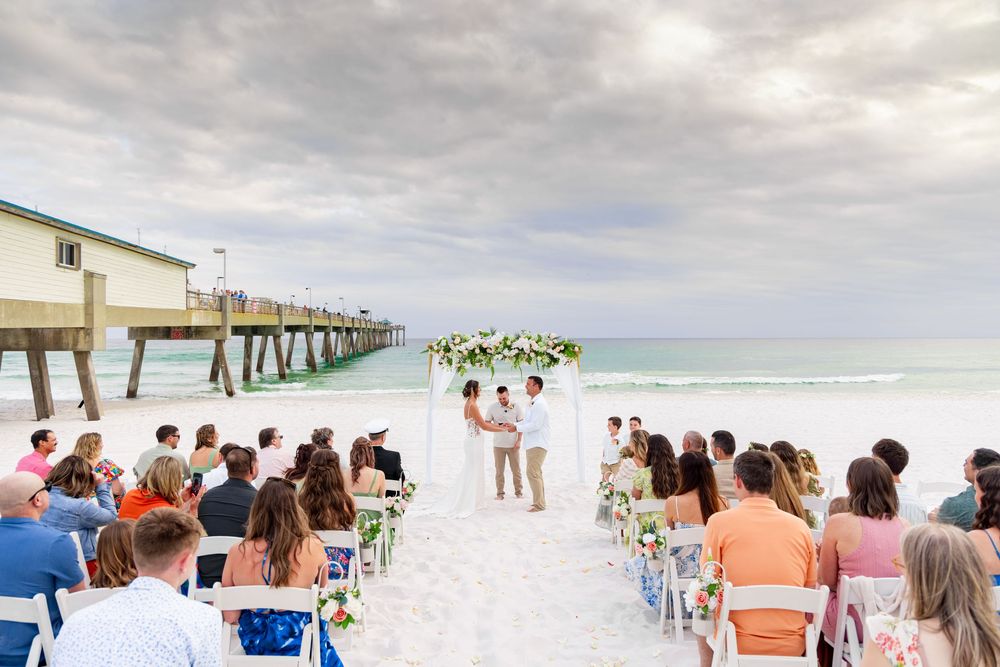 Beach wedding ceremony with floral arch, guests seated on white chairs, ocean waves and wooden pier in background.