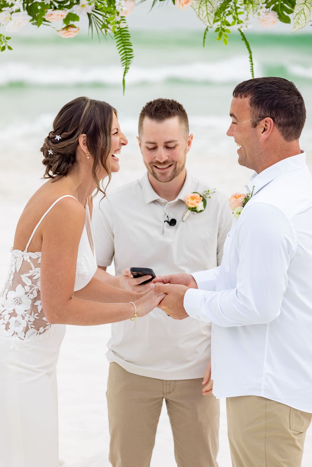 Bride and groom exchange rings during a beach wedding ceremony officiated by a smiling man in a white shirt.