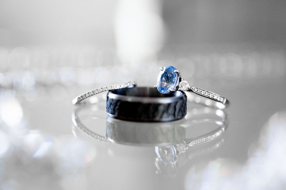 Silver bangle bracelet with diamond accents beside a blue gemstone engagement ring and black wedding band on reflective surface.