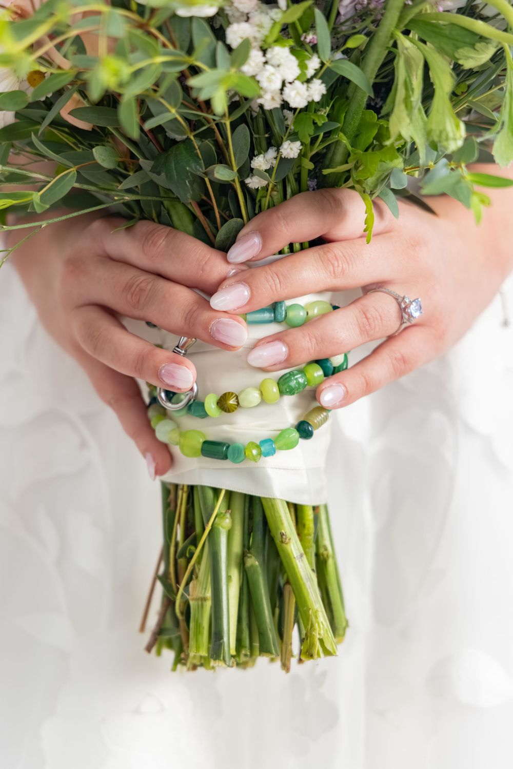Bride holding green floral wedding bouquet with beaded bracelet and diamond ring, featuring elegant nail art.