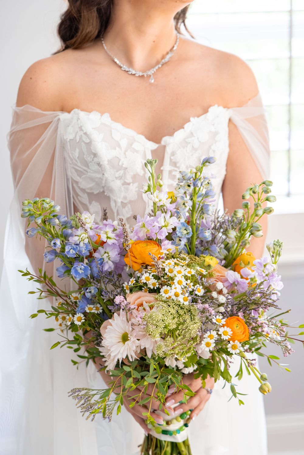 Bride holding a wildflower wedding bouquet with blue, orange, and white blooms in an off-shoulder white gown.