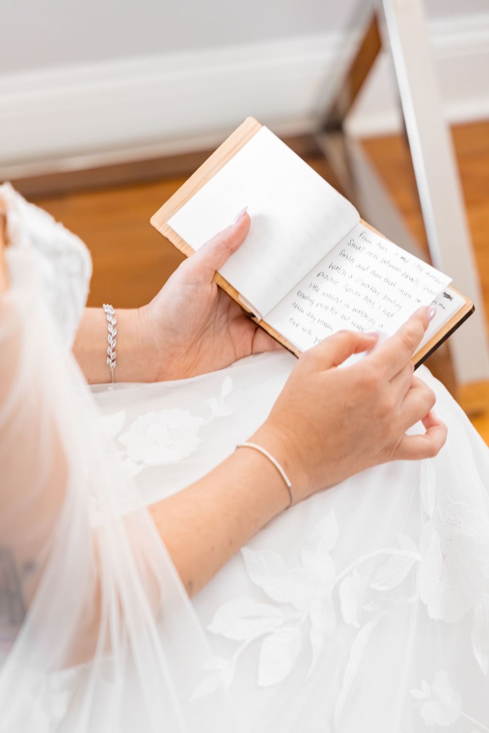 Bride in white lace wedding dress reading vows from a small book while wearing a silver bracelet.