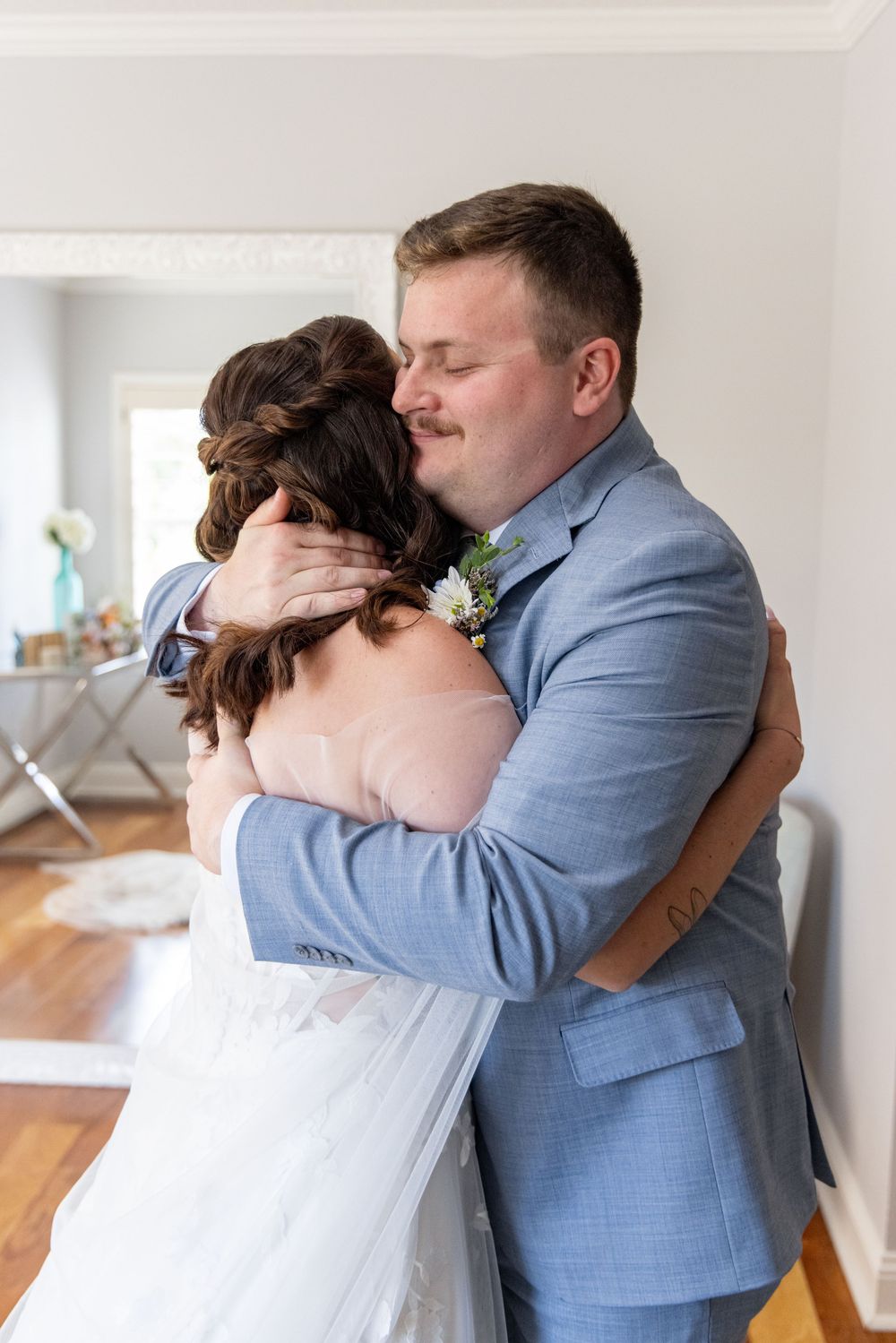 Groom in blue suit embraces bride in emotional first look moment indoors on wedding day.