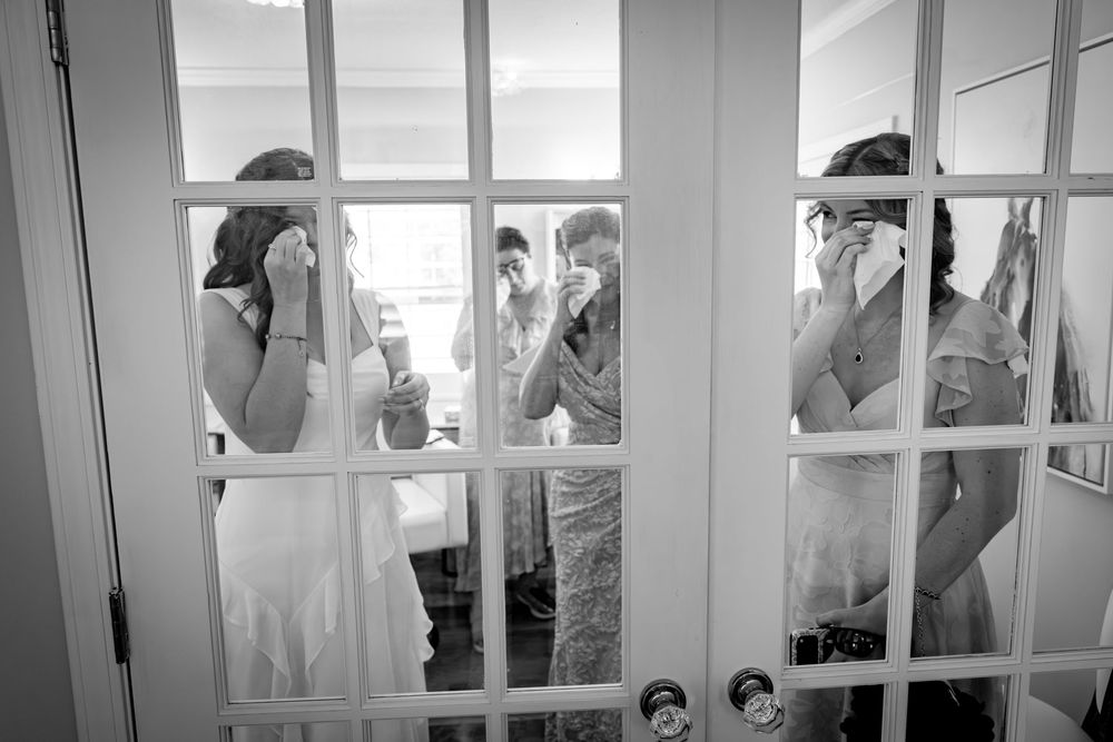 Black and white photo of emotional wedding party crying behind French doors during bridal preparations.
