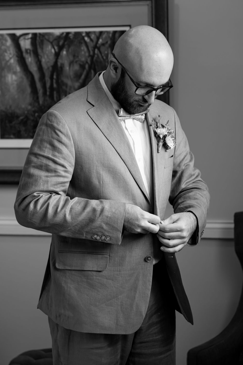 Groom in a tan suit and bow tie buttoning his jacket with a floral boutonniere, black and white photo.