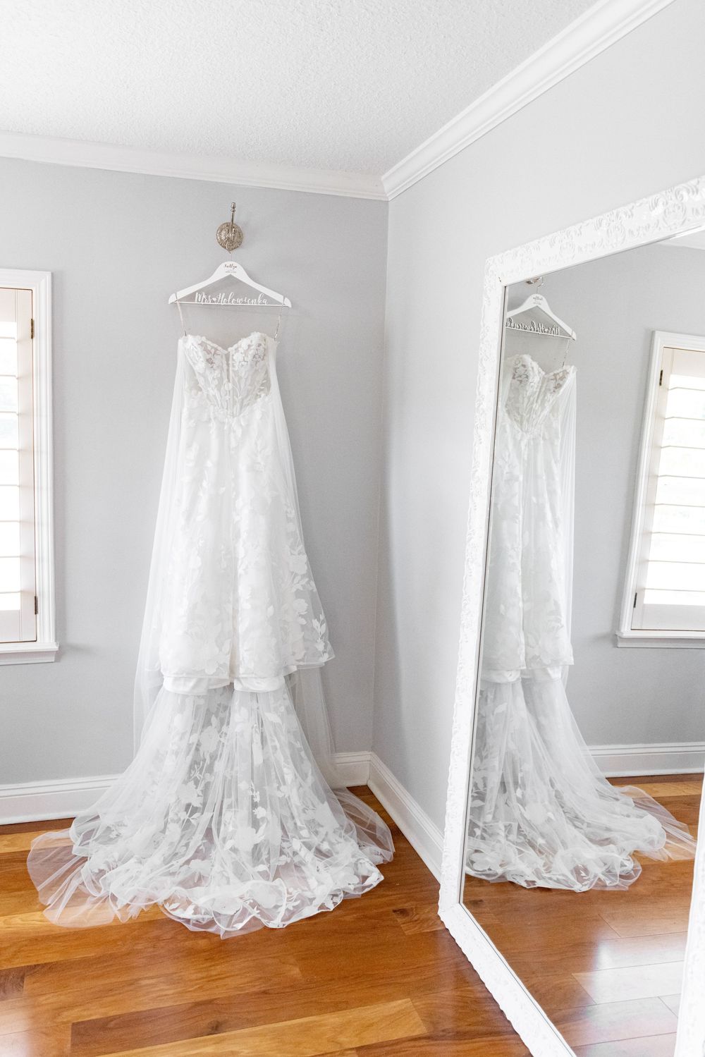 Elegant white lace wedding gown hanging on wall reflected in large mirror on hardwood floors.