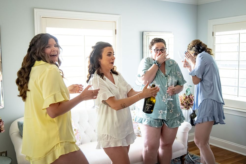 Four bridesmaids in pastel pajamas laughing and celebrating together in a bright room during a fun bridal party getting ready.