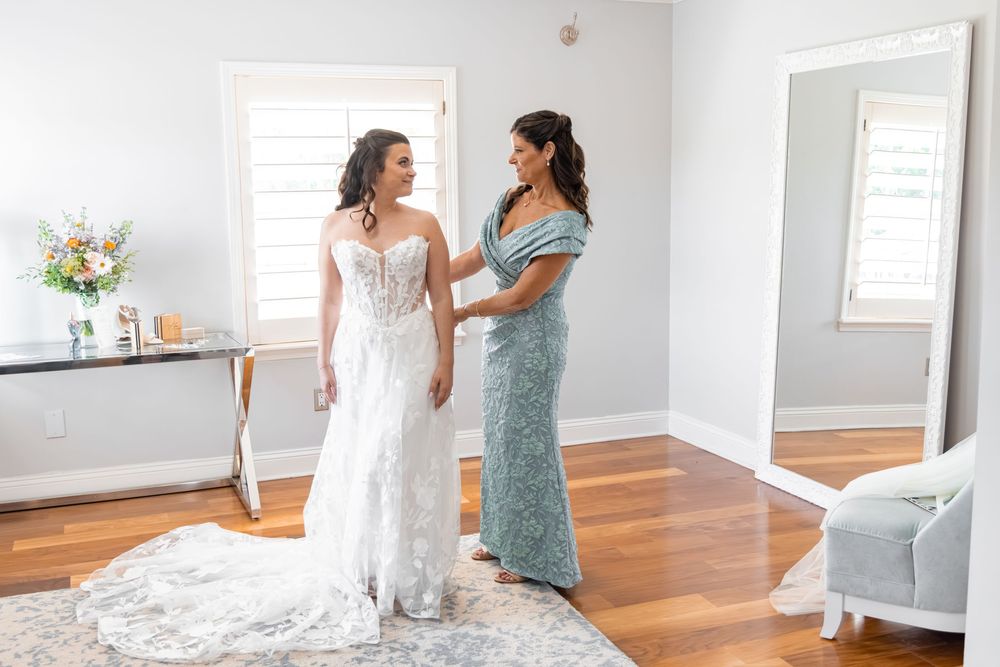 Bride in white lace gown stands with woman in blue dress in bright room with mirror and flowers.