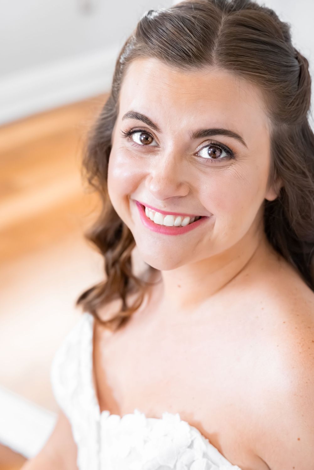 Smiling bride with brown hair and pink lips in a white wedding dress looking up at the camera.