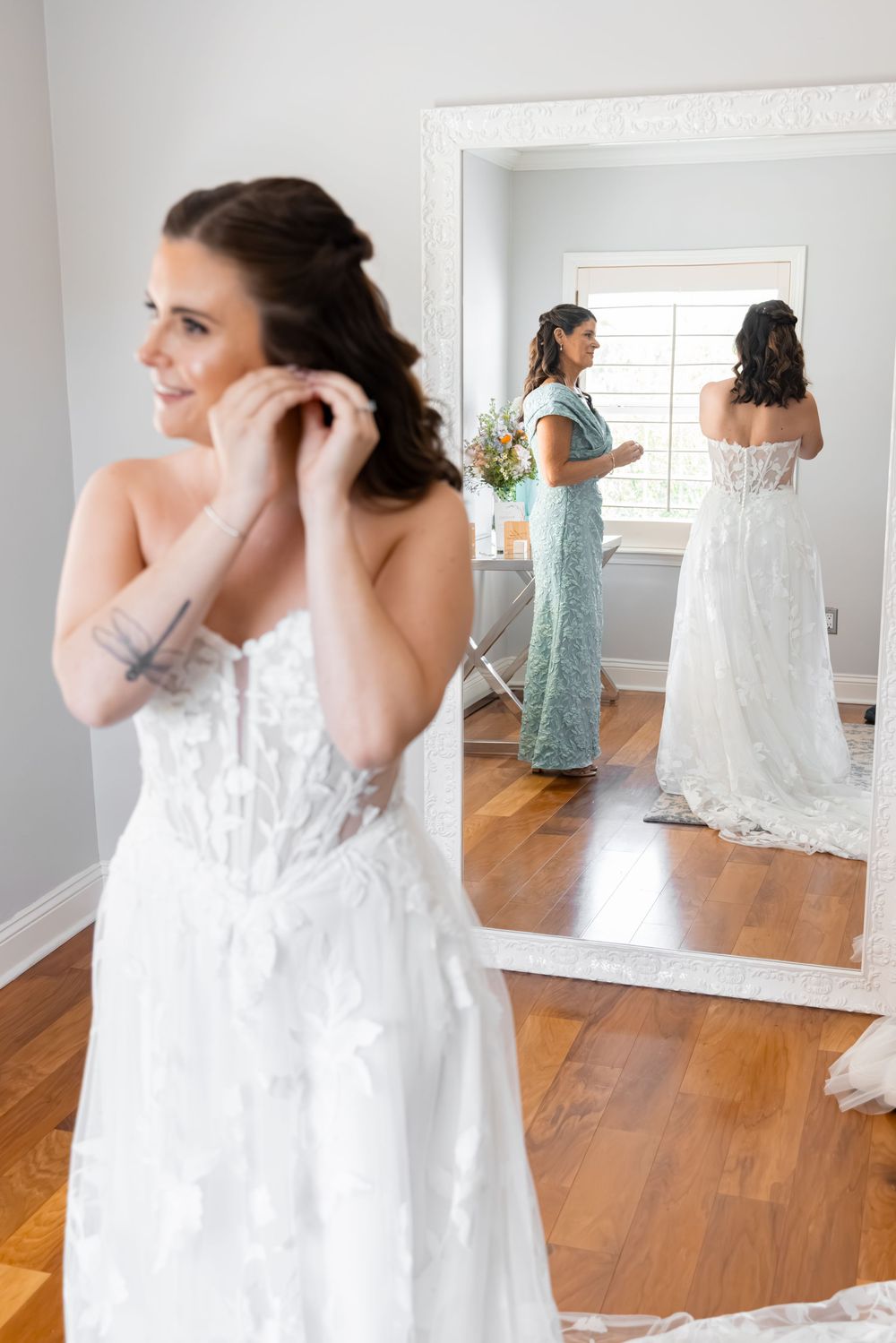 Bride in lace wedding dress adjusting earring while bridesmaid assists in mirror reflection.