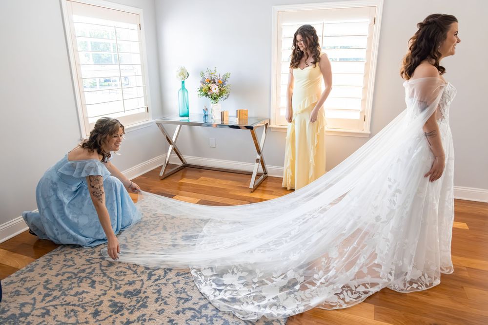 Bridesmaids helping bride with long lace wedding gown train in bright room before wedding ceremony.