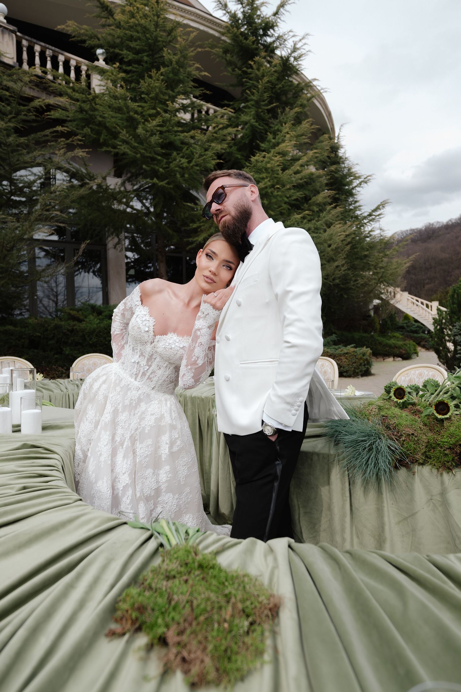 Bride in lace gown leans on groom in white tuxedo at elegant outdoor wedding with green decor.