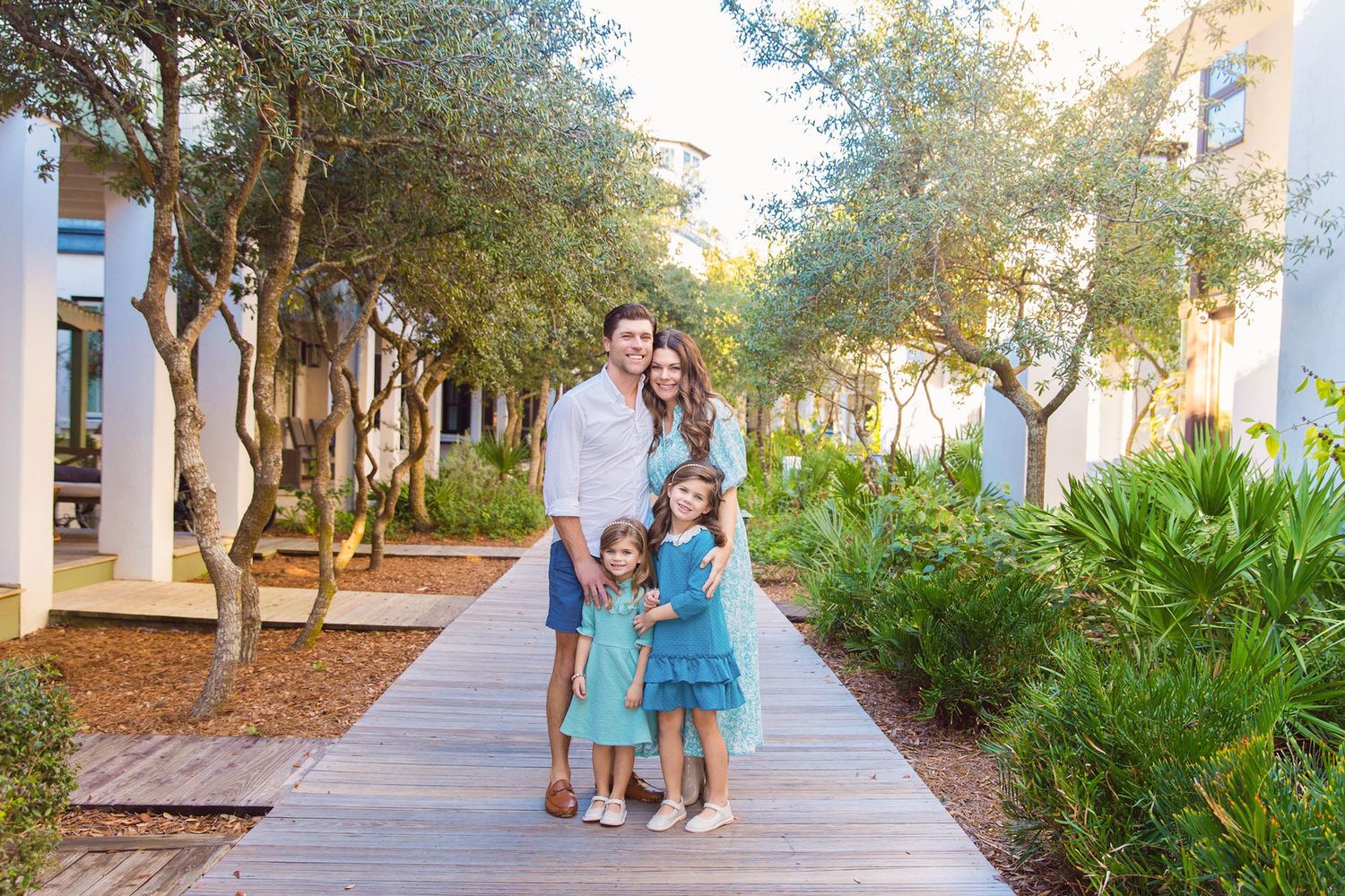 Family of four posing on a wooden boardwalk path surrounded by lush green trees and tropical landscaping.