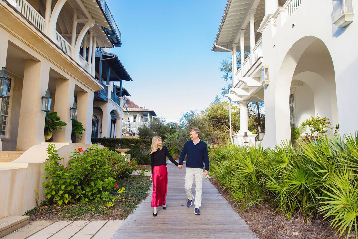 Couple holding hands walking on a wooden boardwalk path between elegant white resort buildings surrounded by tropical landscaping.