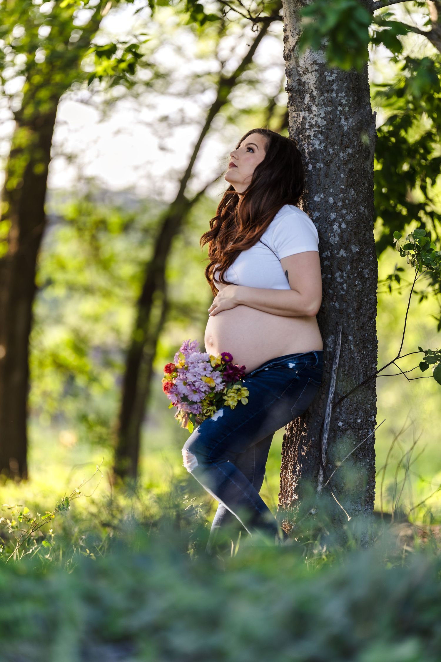 An editorial maternity portrait of a woman leaning against a tree in a sun-drenched field, showcasing the un-staged nuances of her motherhoo