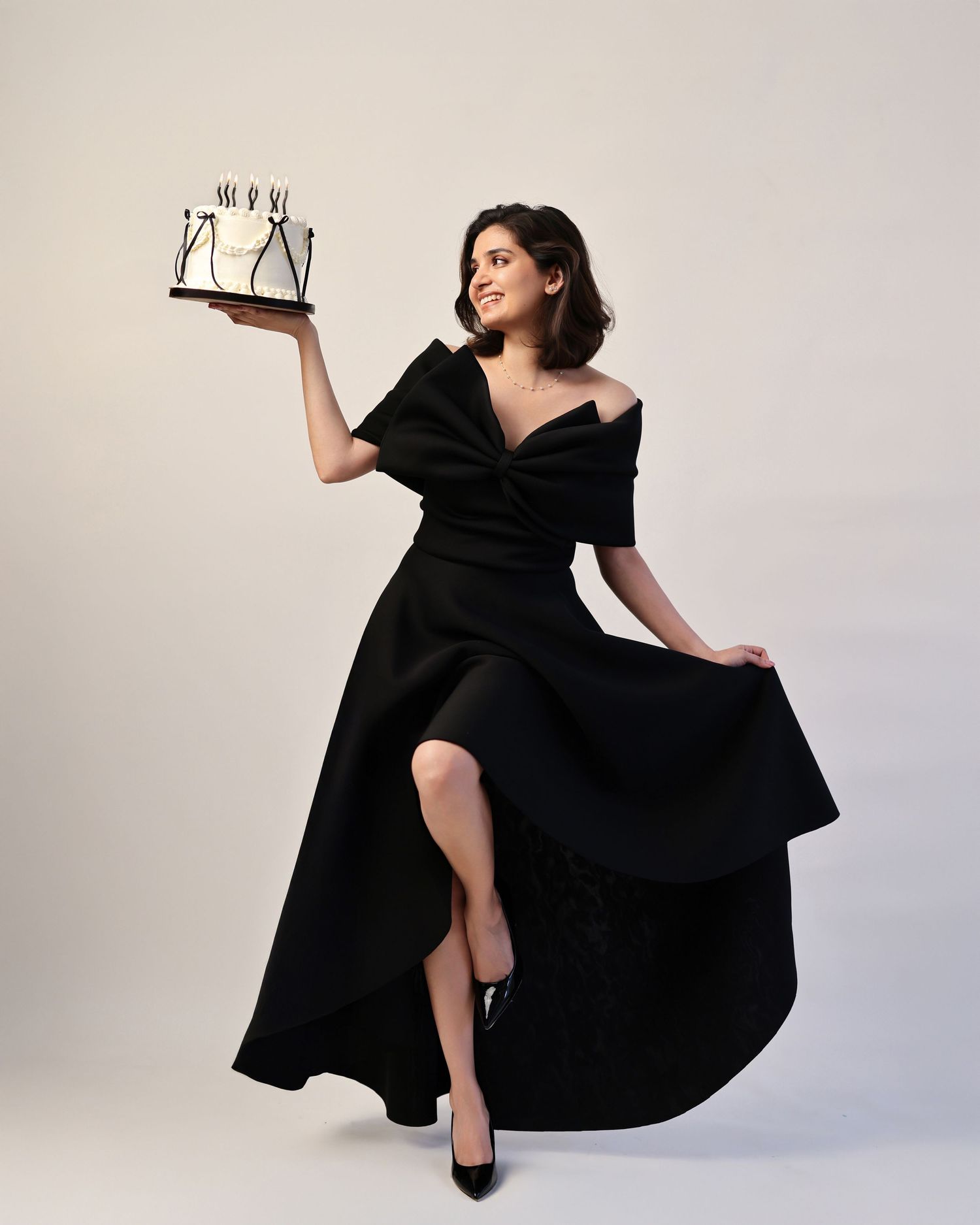 Woman in black dress holding a decorated cake with black bows on wall in dramatic spotlight.