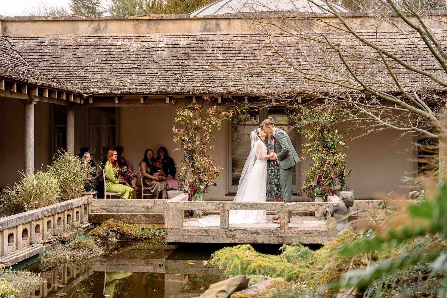 Couple shares a kiss on a wooden bridge over a pond at a Japanese garden wedding venue with guests seated nearby.