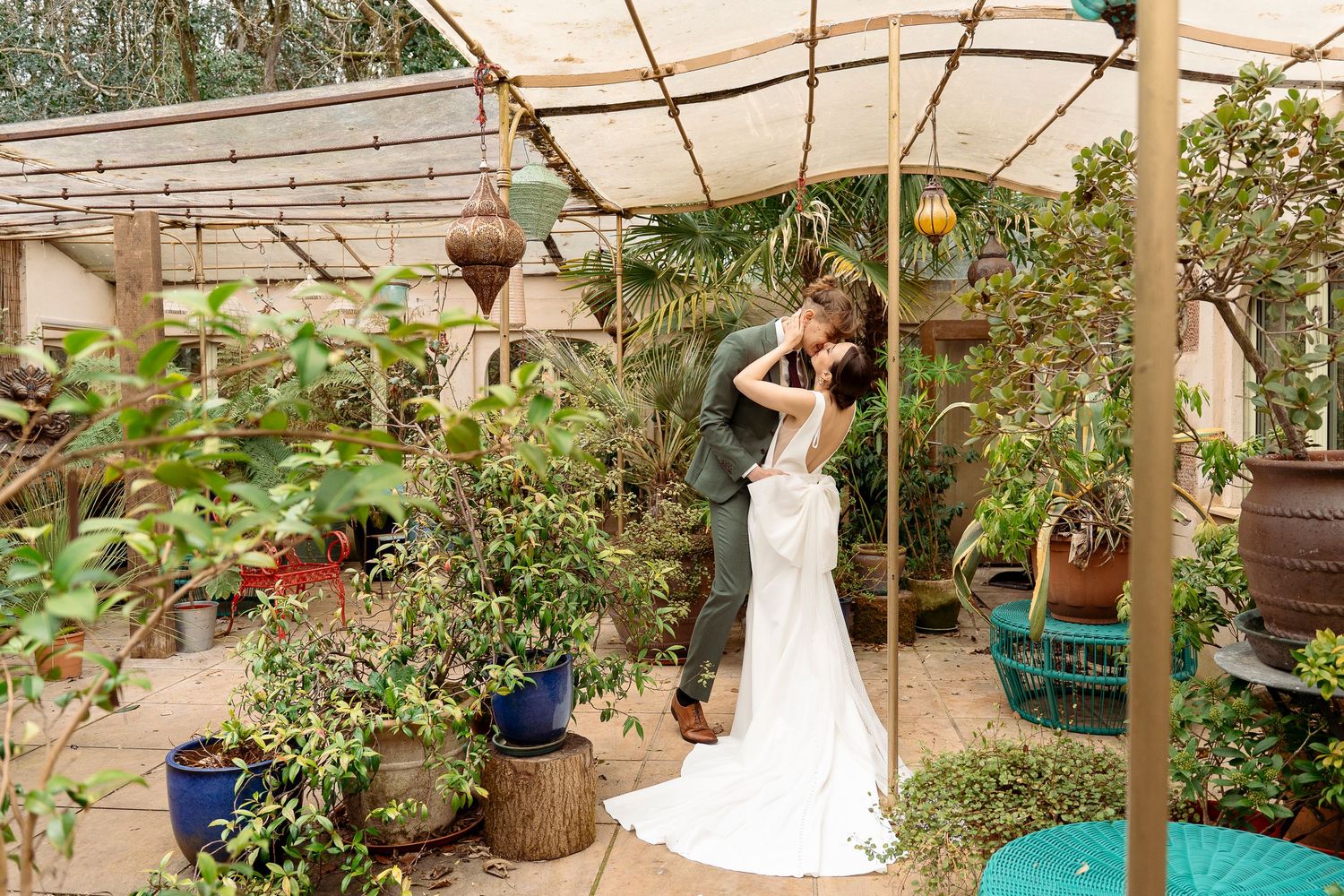 A bride and groom share a romantic kiss surrounded by lush greenery inside a charming greenhouse wedding venue.