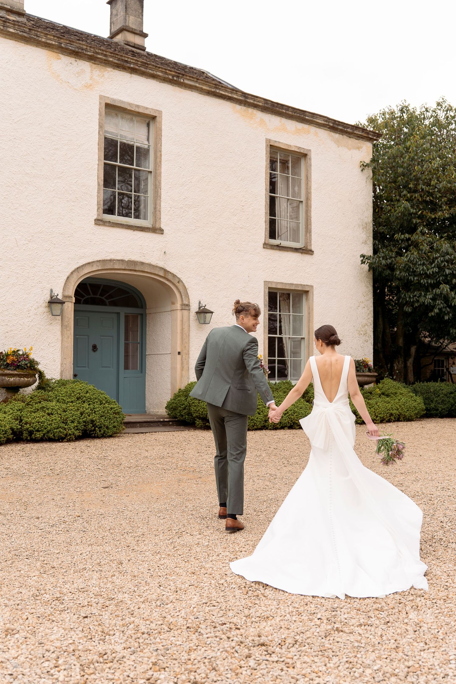 Bride and groom walking hand in hand toward a white country house with a blue door on their wedding day.