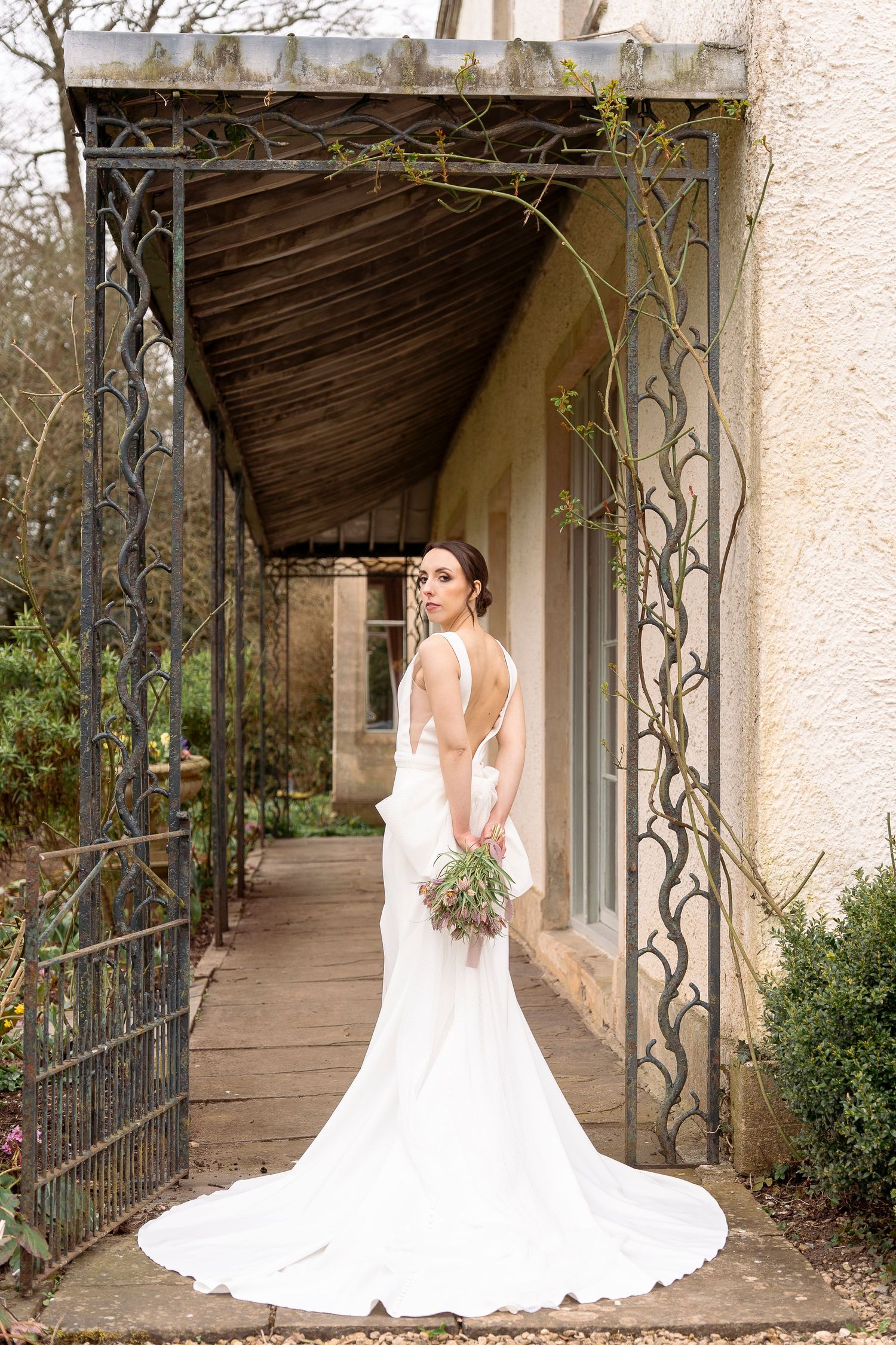 Bride in elegant white backless gown with train holds bouquet under ornate iron pergola at garden venue.