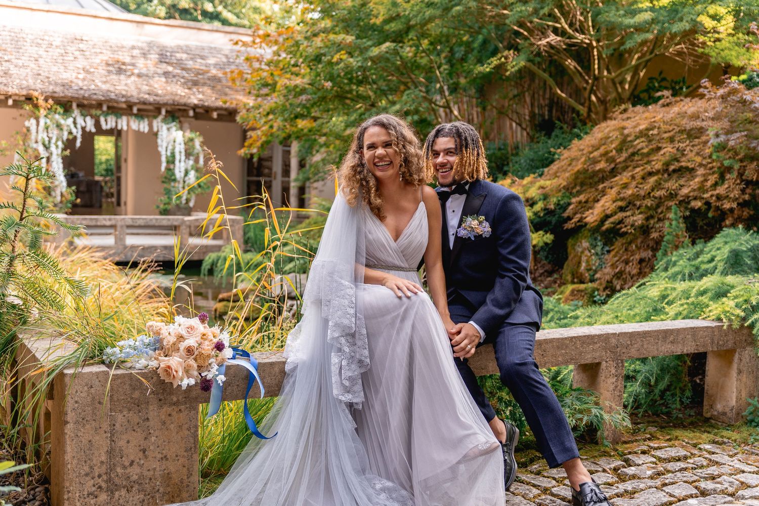 Bride in silver gown and groom in navy suit pose together in a lush garden wedding setting.