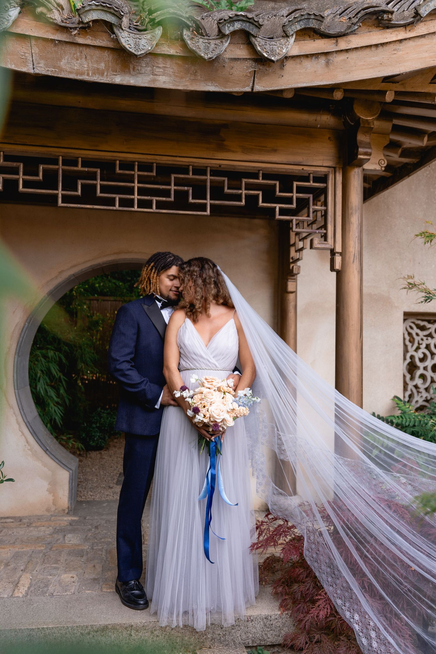 Bride and groom share a romantic moment under a Chinese pavilion, her veil flowing in the breeze.