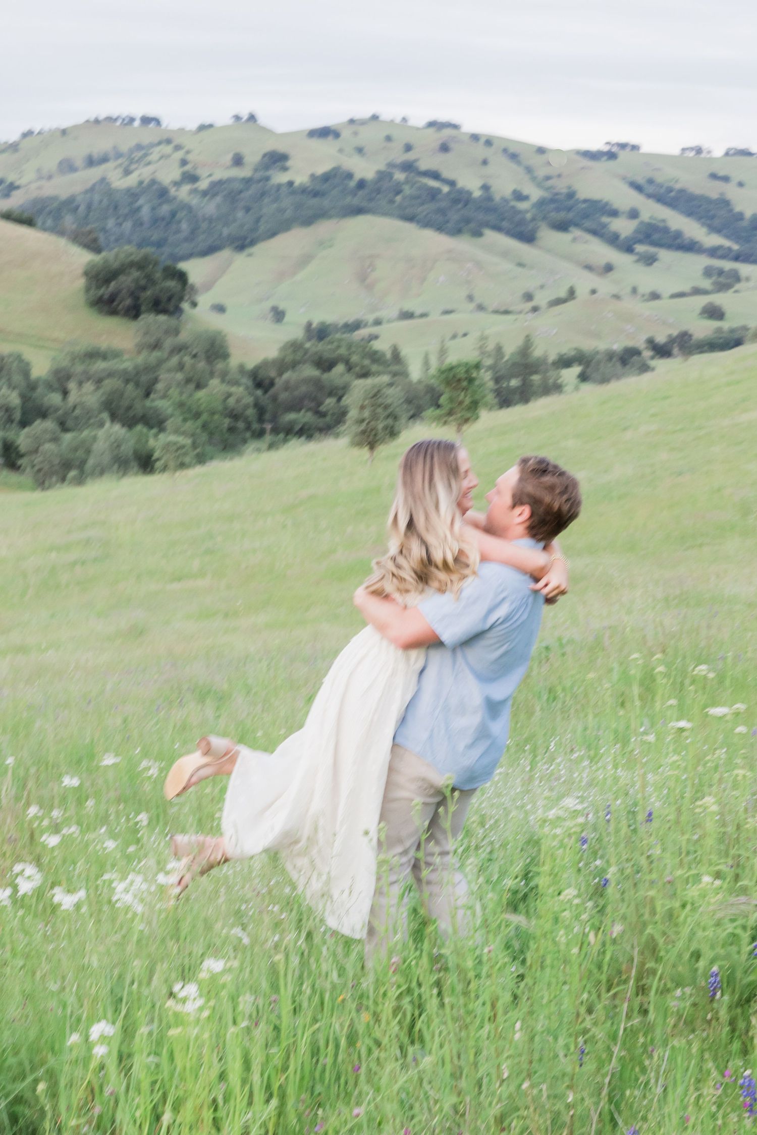 Couple embraces in front of elegant brick building with arched windows and lush greenery at golden hour.