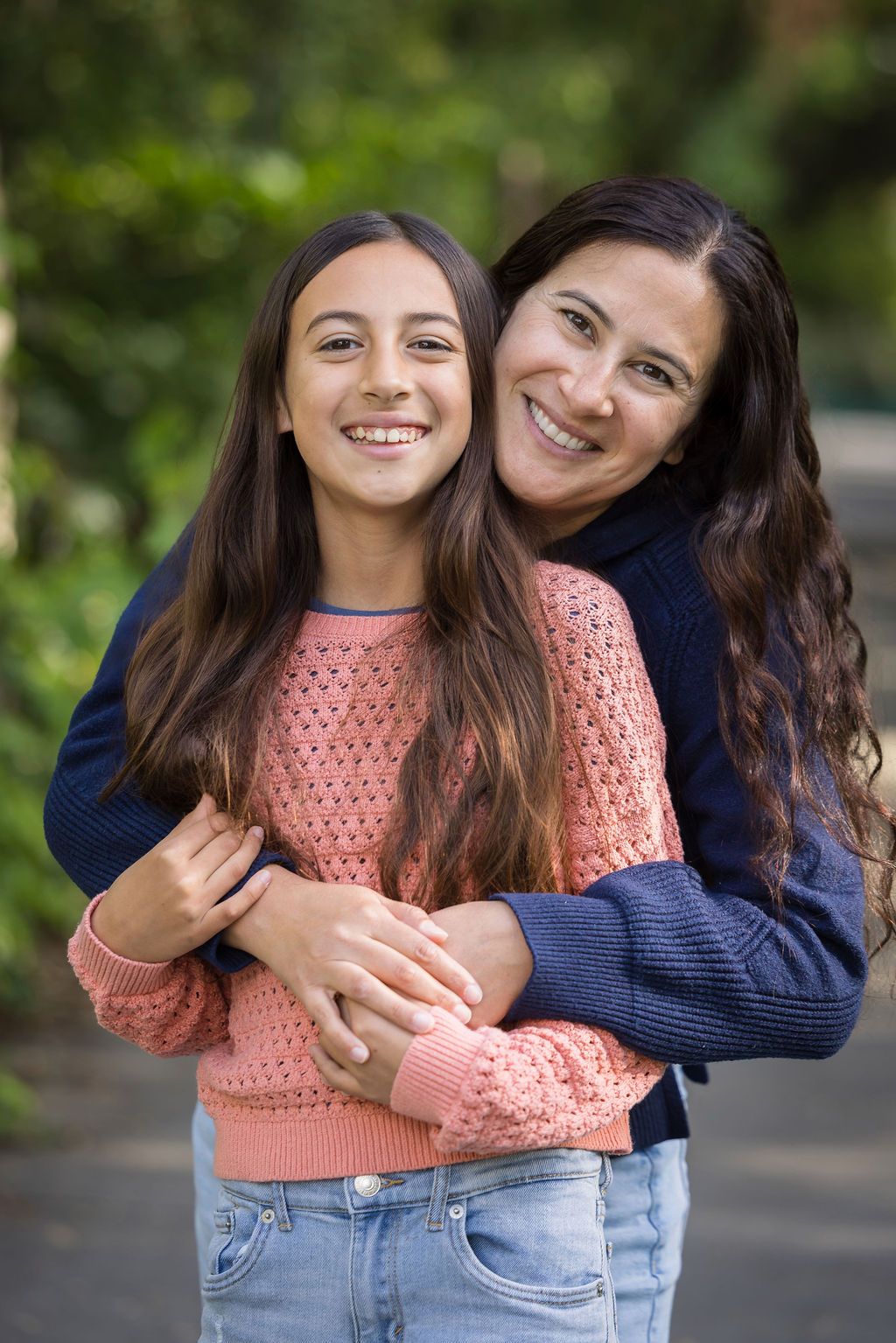 A smiling mother hugs her teenage daughter outdoors, both wearing sweaters, surrounded by lush green foliage.