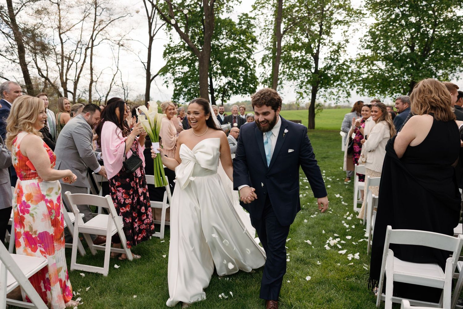 Bride and groom walk down outdoor wedding aisle holding hands, guests celebrating with flower petals on green lawn.
