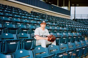 Young baseball pitcher in uniform sits among teal stadium seats holding a glove at an empty ballpark.