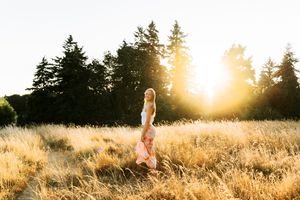 Woman in pink dress walking through golden field at sunset with tall evergreen trees in background.