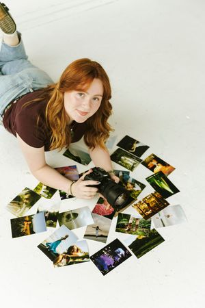 Young redhead woman lying on white floor holding camera surrounded by spread out colorful printed photographs.