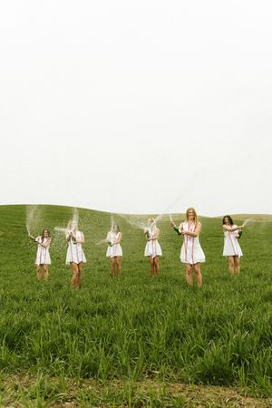 Six women in white dresses celebrate in a green field, popping champagne bottles outdoors.