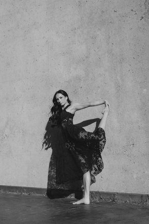 Elegant woman in black lace dress performing a graceful dance pose against a gray wall in black and white photo.