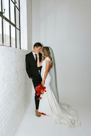 Bride and groom share a romantic moment by a white brick wall, bride holding red floral bouquet in wedding attire.