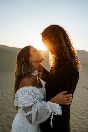 Couple embracing at sunset in desert sand dunes, bride in white lace off-shoulder gown, golden backlight.