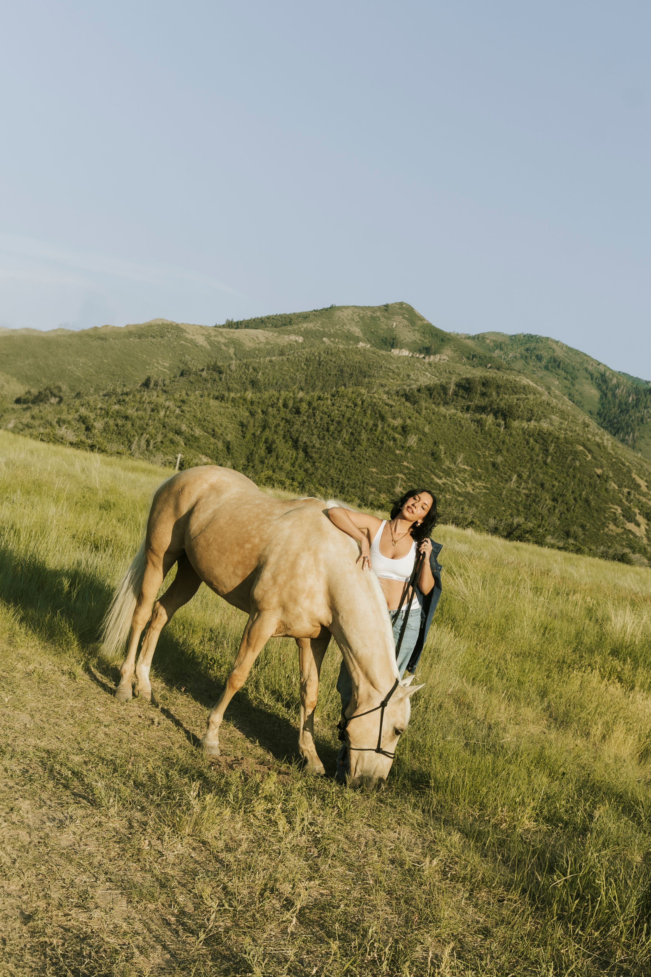 Woman in white top leans affectionately on a palomino horse grazing on a mountain meadow under a clear sky.